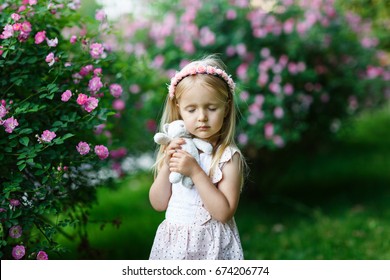 Beautiful little girl presses to the face a teddy bear in the street against a background of blossoming dogrose
