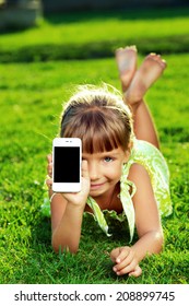 Beautiful Little Girl Is Showing A Phone Touch Screen And Lying On A Green Grass On A Warm Sunny Summer Day.