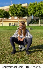 Beautiful Little Girl Sitting On Top Of Her Soccer Ball, Wears A Mask To Protect Herself From The Coronavirus While Doing Sports In A Beautiful Park With Natural Grass. Selective Focus