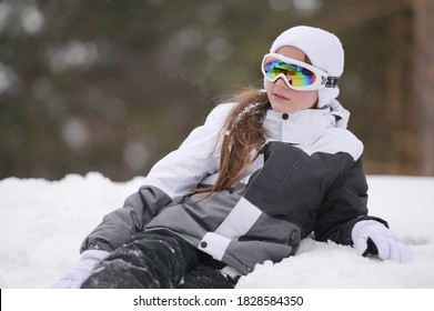 Beautiful Little Girl In Ski Goggles And Suit Jacket With Gloves Lying On White Snow In Winter Park Resort