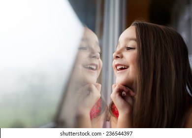 Beautiful Little Girl Smiling And Watching Out The Window. A Child Looks Out The Window. Young Girl Looking From Window. Portrait Of Cheerful Kid Sits At Window.