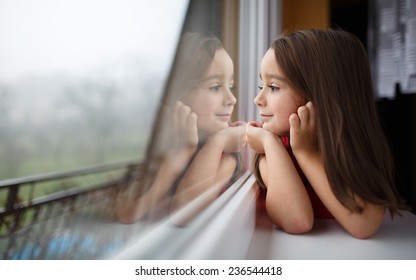 Beautiful Little Girl Smiling And Watching Out The Window. A Child Looks Out The Window. Young Girl Looking From Window. Portrait Of Cheerful Kid Sits At Window.