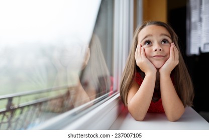 Beautiful Little Girl Smiling And Watching Out The Window. A Child Looks Out The Window. Young Girl Looking From Window. Portrait Of Cheerful Kid Lies At Windowsill.