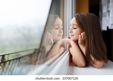 Beautiful Little Girl Smiling And Watching Out The Window. A Child Looks Out The Window. Young Girl Looking From Window. Portrait Of Cheerful Kid Lies At Windowsill.