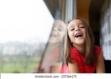 Beautiful Little Girl Smiling And Watching Out The Window. A Child Looks Out The Window. Young Girl Looking From Window. Portrait Of Cheerful Kid Sits At Windowsill.