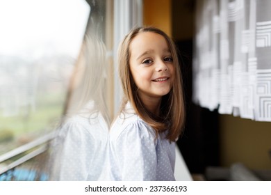 Beautiful Little Girl Smiling And Watching Out The Window. A Child Looks Out The Window. Young Girl Looking From Window. Portrait Of Cheerful Kid Sits At Windowsill.