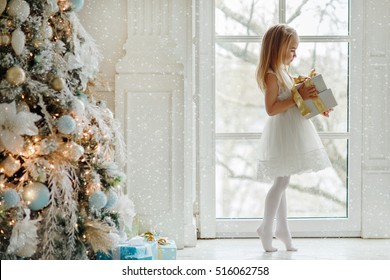 Beautiful little girl standing on tiptoes at the large window and holding a gift, in the bright New Year's interior