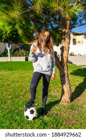 Beautiful Little Girl Stepping On A Soccer Ball With Her Thumb Up, Wears A Mask To Protect Herself From The Coronavirus While Doing Sports In A Beautiful Park With Natural Grass. Healthy Life Concept