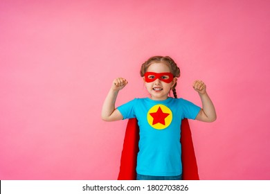 Beautiful Little Girl In A Superhero Costume, In A Red Cape And Mask Shows How Strong She Is, Isolated Against A Pink Background. Cute Kid Playing Superhero. The Concept Of Power Girl. Copy Space.