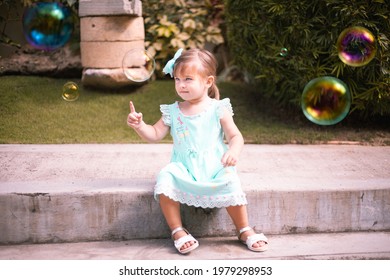 Beautiful little girl toddler in dress playing with bubbles, popping the bubbles in summer time in the garden