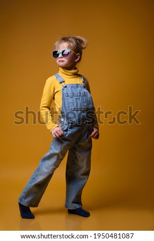 Beautiful little girl wearing stylish denim overall, yellow roll neck jumper and colorful sunglasses standing on bright yellow background. Kids fashion concept