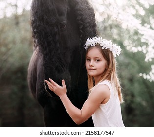 Beautiful little girl with white hair in a spring wreath in the garden with a friesian horse