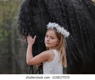 Beautiful little girl with white hair in a spring wreath in the garden with a friesian horse