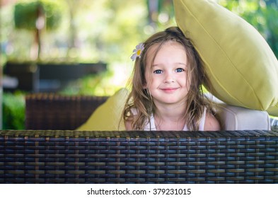 Beautiful Little Girl In A White Sundress Looks Out From Under The Light Green Cushions On Wicker White Sofa.  Sofa Is Outdoors, In The Background Sunlight, Flowers And Green Shrubs.