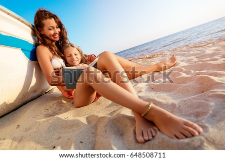 Beautiful little girl and young woman reading a book on the sandy beach. She is sitting next to boat with smile on her face.