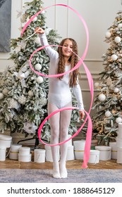 A Beautiful Little Gymnast In A White Sports Dress Doing Rhythmic Gymnastics Exercises Spirals With An Artistic Ribbon In A Fitness Class During The Christmas Season. Sports, Training, Stretching