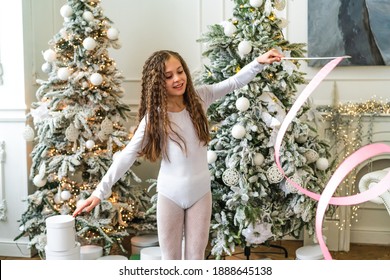 A Beautiful Little Gymnast In A White Sports Dress Doing Rhythmic Gymnastics Exercises Spirals With An Artistic Ribbon In A Fitness Class During The Christmas Season. Sports, Training, Stretching