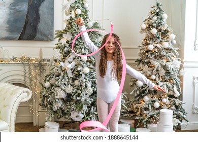 A Beautiful Little Gymnast In A White Sports Dress Doing Rhythmic Gymnastics Exercises Spirals With An Artistic Ribbon In A Fitness Class During The Christmas Season. Sports, Training, Stretching