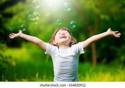 Beautiful little haired hair girl, has happy fun smiling face, pretty eyes, short hair, playing soap bubbles, dressed in white t-shirt. Child portrait. Creative concept. 