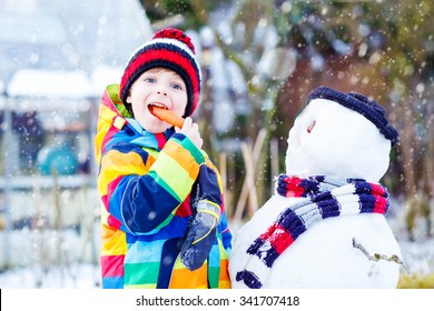 Beautiful Little Kid Boy Making A Snowman And Eating Carrot. Child Playing And Having Fun With Snow On Cold Day. Active Outdoors Leisure With Kids In Winter.