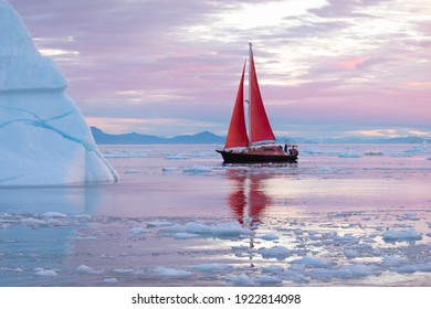 Beautiful little red sailboat in the arctic next to a massive iceberg showing the scale in pink dawn light. Ilulissat, Disko Bay, Greenland.
