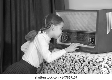 A beautiful little schoolgirl girl in a white blouse and black long skirt, with neatly braided pigtails on her head.She presses the keys of the old radio. Interior of the fifties of the last century.