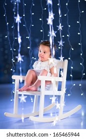 Beautiful little toddler girl with curly hair wearing a white dress sitting in a white rocking chair in a dark room with Christmas lights