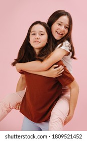Beautiful little twin sisters happily playing together, one riding the other. Over pink background, studio shot. They wear pink and blue pants in contrast with their shirts.
