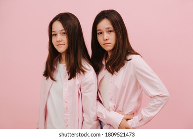 Beautiful Little Twin Sisters Standing One Behind The Other. Over Pink Background, Studio Shot. One Is Smiling, Other Is Not. Both Looking At The Camera.