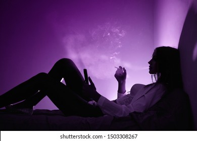 Beautiful lonely girl with smartphone smoking cigarette lying in the bed in dark room