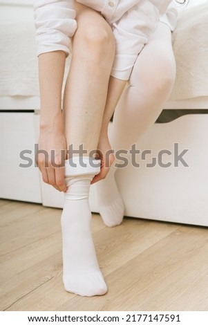 Beautiful long female legs in stockings. Girl putting on stockings at home in a white room. White hosiery. Varicose veins prevention. Woman body in underwear.