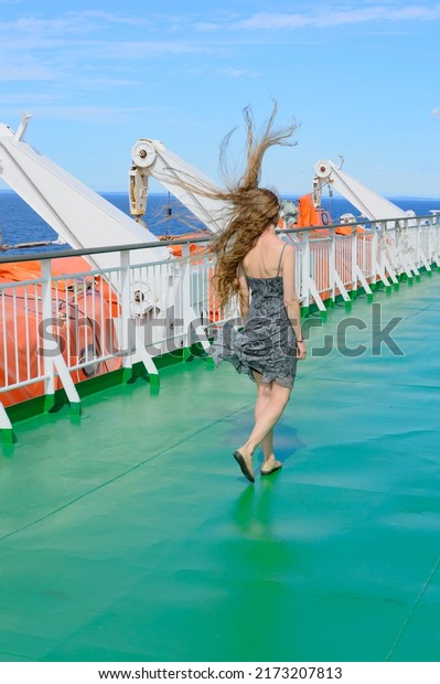 Beautiful long haired blonde girl seen from behind\
dressed in a grey summer dress,  walks along the upper deck of a\
cruise liner. Light blue sky, blue sea, green ship deck floor,\
orange life boats