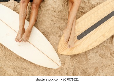 Beautiful long legs of two young girl in sexy bikini lycra on a sand beach after surfing with wooden surf white surfboard board at sunrise sunset. Vacation concept. Summer holidays. Tourism, sport.