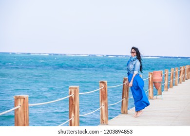 A beautiful long  haired girl in long blue skirt is standing wooden pier against the background the sea   sky
