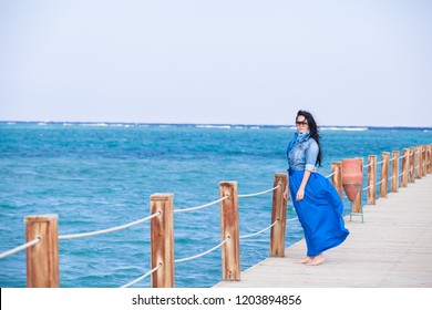 A beautiful long  haired girl in long blue skirt is standing wooden pier against the background the sea   sky
