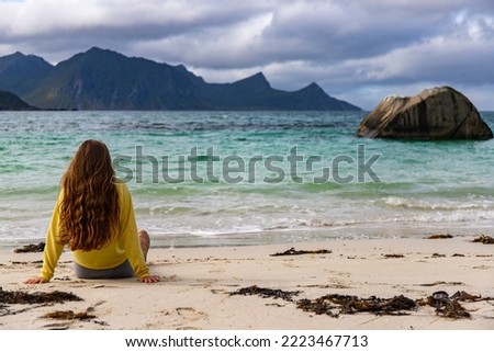 a beautiful long-haired girl sunbathes while sitting on the sand on the famous haukland beach on the lofoten islands in northern norway; relaxing on the beach in the norwegian fjords