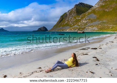a beautiful long-haired girl sunbathes while sitting on the sand on the famous haukland beach on the lofoten islands in northern norway; relaxing on the beach in the norwegian fjords