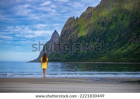 beautiful long-haired girl in a yellow dress walks on the famous beach with the mighty mountains in the background ersfjordstranda in norway, senja, holidays in the norwegian fjords
