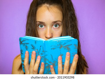 A beautiful long-haired grey-eyed brunette student girl is holding a blue book, a notebook, covering face with it. Young woman studying, reading, preparing for a session. Surprise, consternation. - Shutterstock ID 2011903298