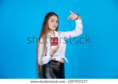 Beautiful long-haired lady in shirt pointing at wee gift box, while looking at camera. Portrait of smiling woman gesturing, while showing decorated red present, isolated on blue. Concept of gesturing.