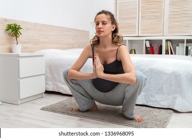 Beautiful long-haired pregnant woman with a big tummy doing a gymnastic exercise and crouching in a yoga pose on the bedside rug and meditating with a smile and eyes closed with palms together.