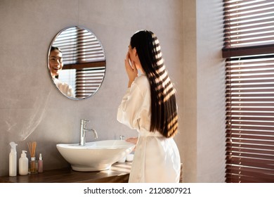 Beautiful long-haired young japanese lady in white bathrobe looking at mirror and touching her face, applying hydrating cosmetics on her skin and smiling, modern bathroom interior, copy space