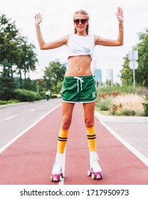 Beautiful Long-legged Girl Posing On A Vintage Roller Skates In Geen Shorts, Knee Socks And White T-shirt In The Skate Park On A Warm Summer Evening. Rollers Quads Derby. Hands Up.