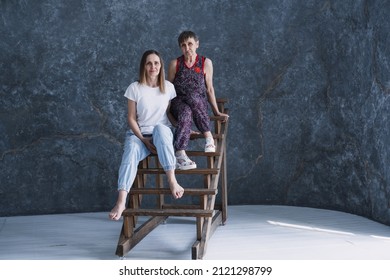 Beautiful Looking Adult Daughter 50 Years Old And Mother Elderly Woman 70 Years Old Sitting Together On Wooden Stairs. Sports Lifestyle