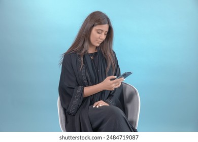 Beautiful looking woman wearing abaya using her mobile phone on solid background. Happy attractive Emirati or saudi lady on blue background sitting - Powered by Shutterstock