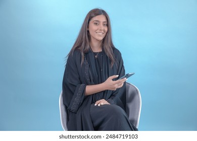 Beautiful looking woman wearing abaya using her mobile phone on solid background. Happy attractive Emirati or saudi lady on blue background sitting - Powered by Shutterstock