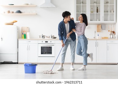 Beautiful loving asian young couple mopping floor in kitchen together, full length photo, copy space. Romantic cheerful chinese lady hugging her smiling boyfriend while cleaning house