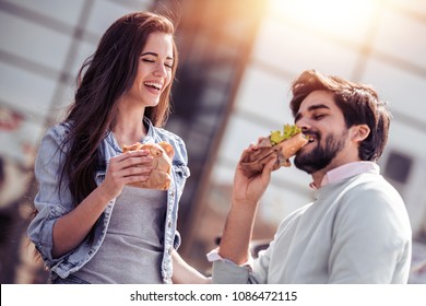 Beautiful loving couple sitting on the bench and eating sandwich outdoors.