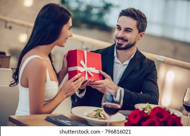 Beautiful Loving Couple Is Spending Time Together In Modern Restaurant. Attractive Young Woman In Dress And Handsome Man In Suit Are Having Romantic Dinner. Celebrating Saint Valentine's Day.