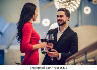 Beautiful Loving Couple Is Spending Time Together In Modern Restaurant. Attractive Young Woman In Dress And Handsome Man In Suit Are Having Romantic Dinner. Celebrating Saint Valentine's Day.